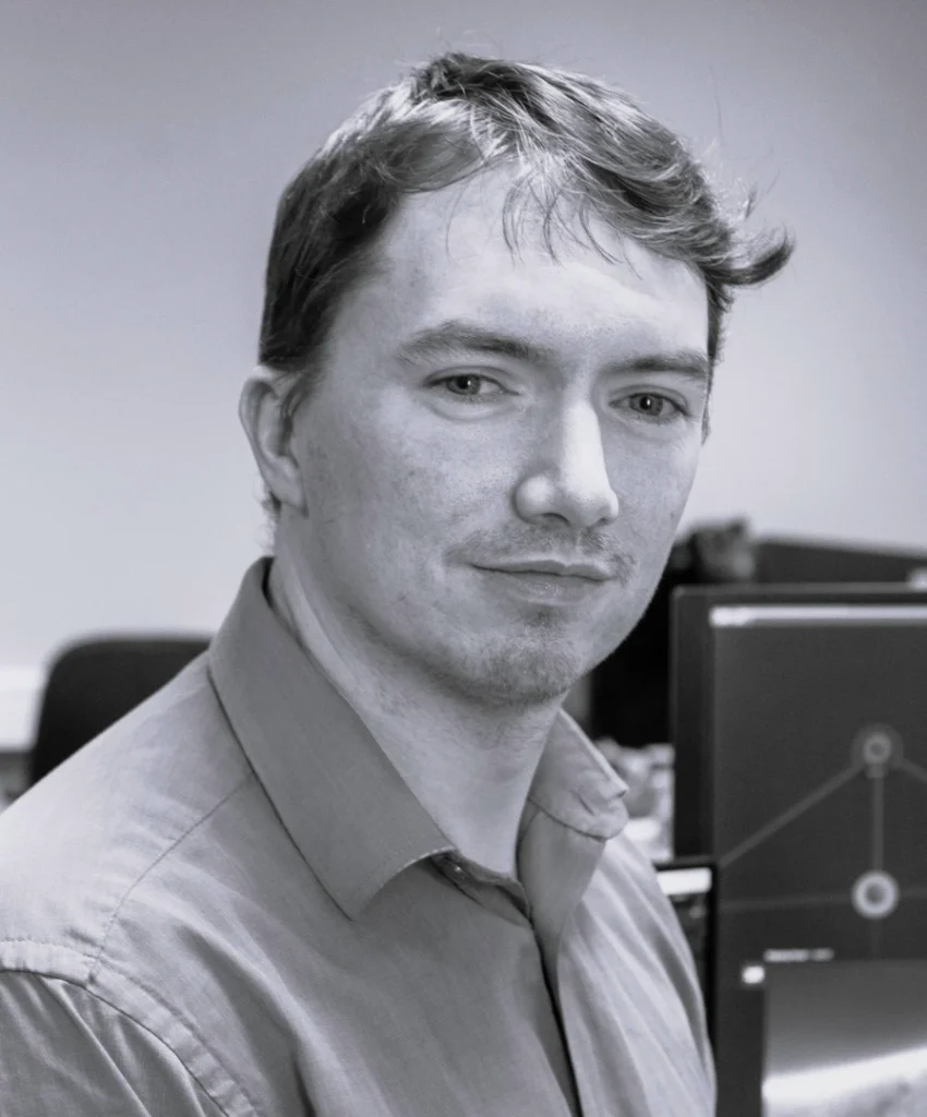 Black and white photograph of a man wearing a collared shirt in an office setting with computer monitors displaying graphs and charts in the background. The scene suggests a professional environment focused on data analysis or financial work.