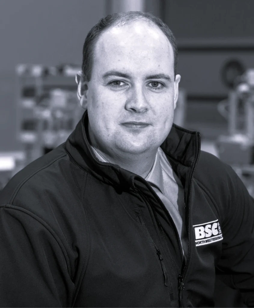 Photograph of a person wearing a black jacket with a "BSC Northwest Region" logo on the chest, seated indoors with industrial or workshop equipment blurred in the background. The grayscale image focuses on the upper body and clothing details, with the face obscured.