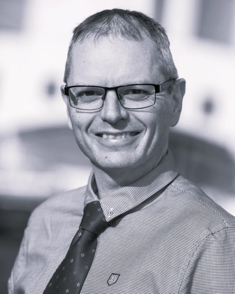 Black and white photograph of a man wearing a checkered shirt and patterned tie, with a blurred background suggesting an indoor setting. The face is obscured by a grey square, focusing attention on clothing and posture rather than identity.