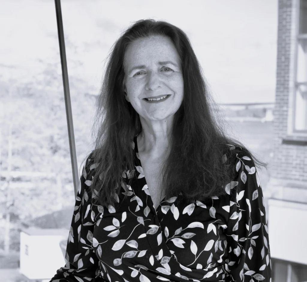 Black and white photograph of a person with long hair wearing a patterned shirt with leaf designs, standing indoors near a window. Background shows blurred outdoor scenery including trees and a building.