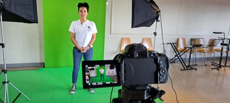 Photograph of a person standing in front of a green screen setup in a studio with professional lighting and a camera on a tripod capturing the scene. The studio includes chairs and music stands in the background, indicating a multi-purpose room used for recording or photography sessions.