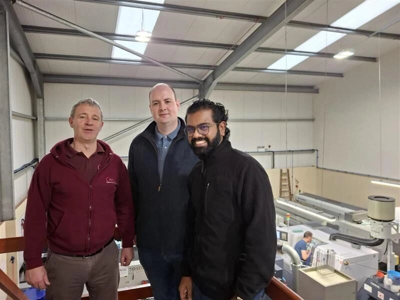 Photograph showing three men standing inside an industrial workshop with machinery and a worker operating equipment in the background. The men wear casual work attire, including a maroon zip-up jacket, a dark blue jacket, and a black jacket, with the workshop featuring high ceilings and bright lighting.