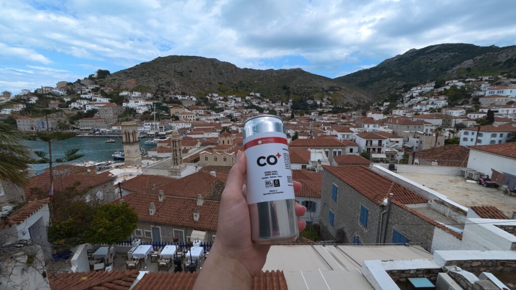 Photograph shows a hand holding a silver CO₂ canister above a coastal hillside town with red-tiled roofs, harbor, and rugged mountains. Canister label provides focal point, while blue water, white buildings, church tower, and partly cloudy sky establish a scenic Mediterranean setting.