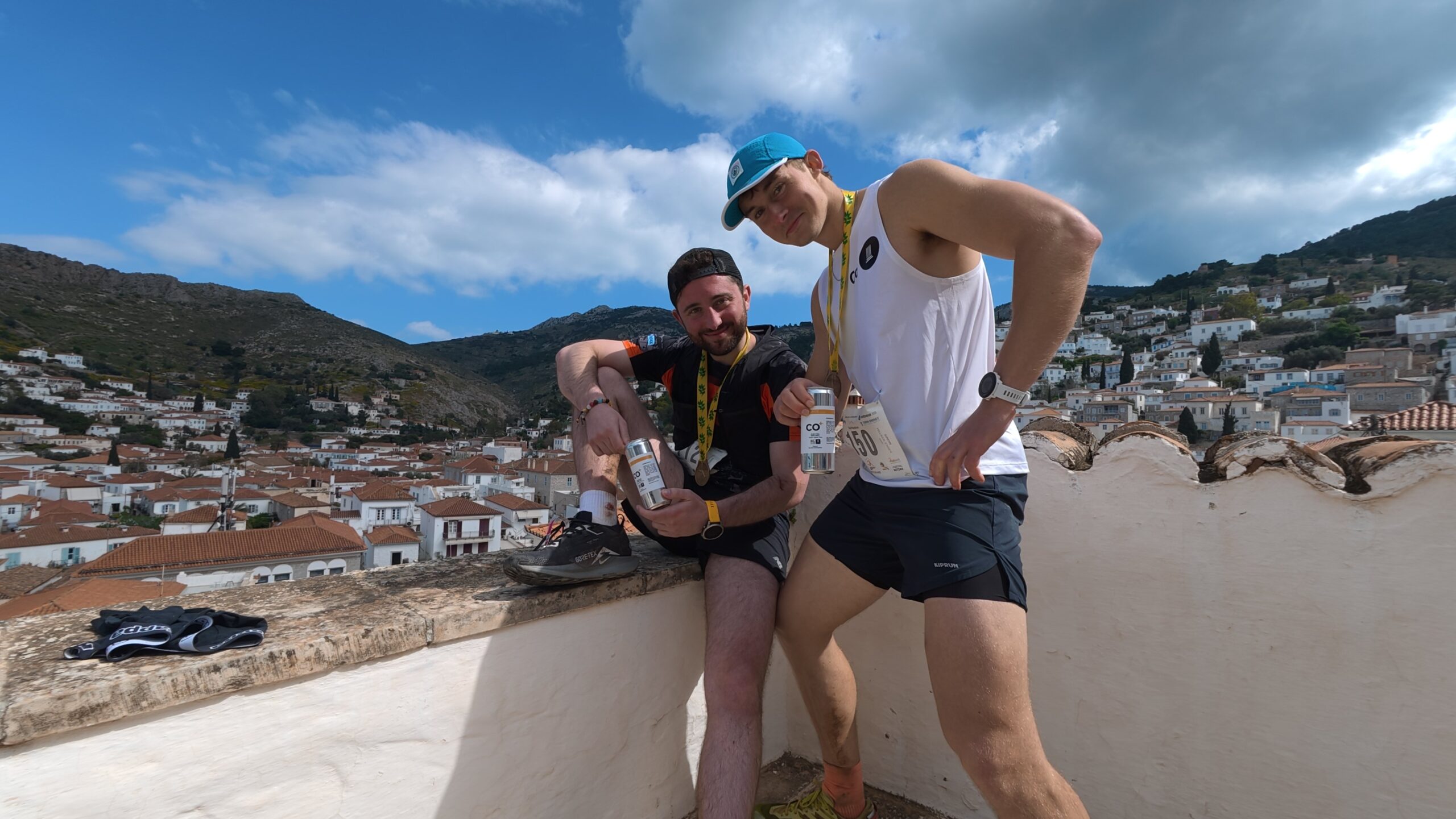 Photograph shows two men posing on a rooftop terrace overlooking a hillside town and rugged mountains under a partly cloudy blue sky. One man wears a white sleeveless shirt and teal cap, while the other wears black athletic clothing and holds a bottle, with stone arches and white buildings visible in background.