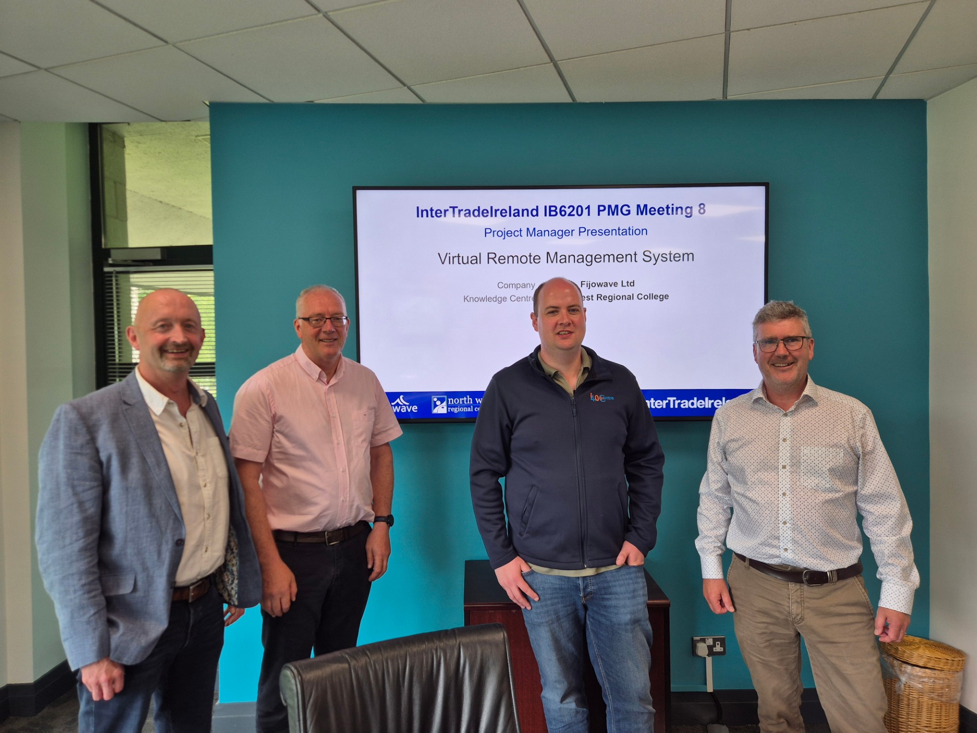 Photograph shows four men standing at a project meeting presentation. Television screen displays project-management text and partner logos, with men posed in front of a teal wall inside an office.